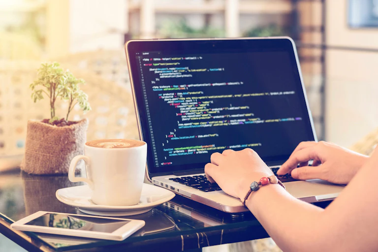 A photo of a person's hands typing on a laptop showing computer code, with a phone and cup of coffee on the table.