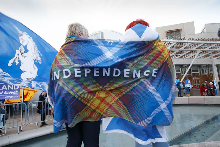 View from behind of two people wrapped in a flag blending tartan and the Scotland flag and reading INDEPENDENCE