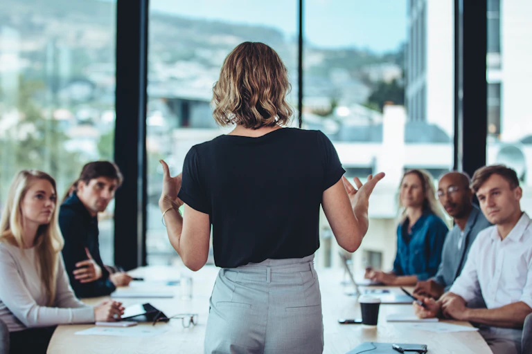 Women in black top holding a meeting, standing in front of seated colleagues, city and mountains background