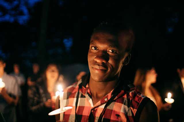 A young man in a plaid shirt holds up a candle against a night sky
