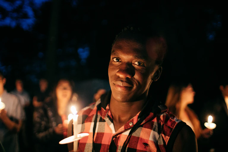 A young man in a plaid shirt holds up a candle against a night sky