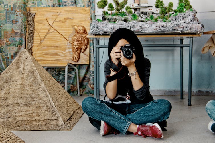 A young woman in a hijab and jeans sits on the floor taking a photograph.