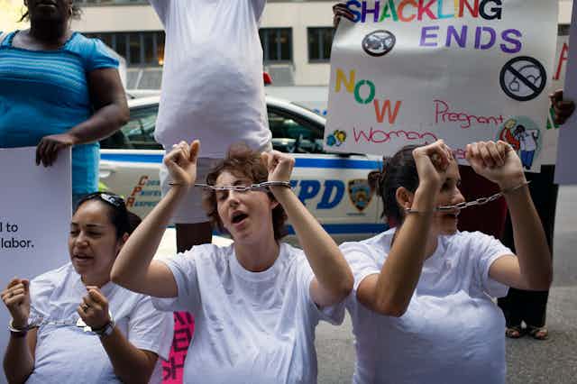 Women wear white shirts and raise their hands with hand cuffs, in front of a sign that says ' Shackling ends now'