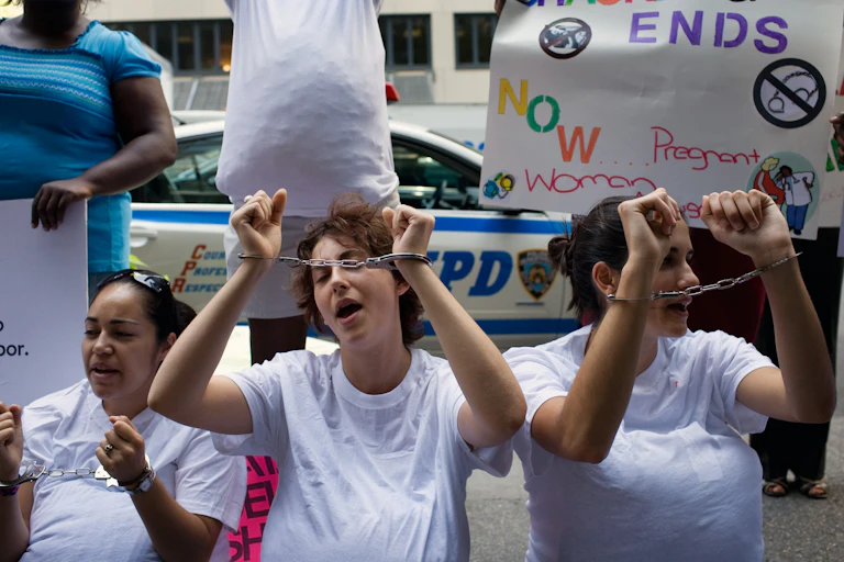 Women wear white shirts and raise their hands with hand cuffs, in front of a sign that says ' Shackling ends now'