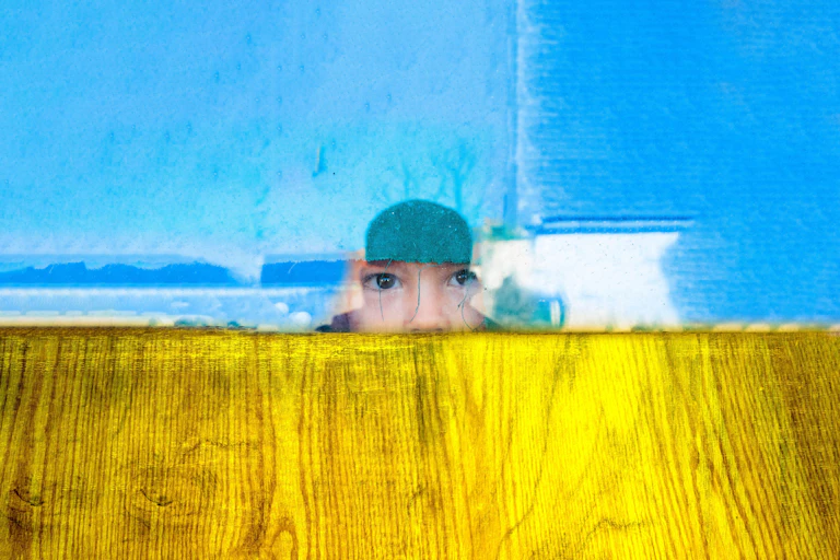 A child looking out of a broken window amid colours reminiscent of the Ukrainian flag.