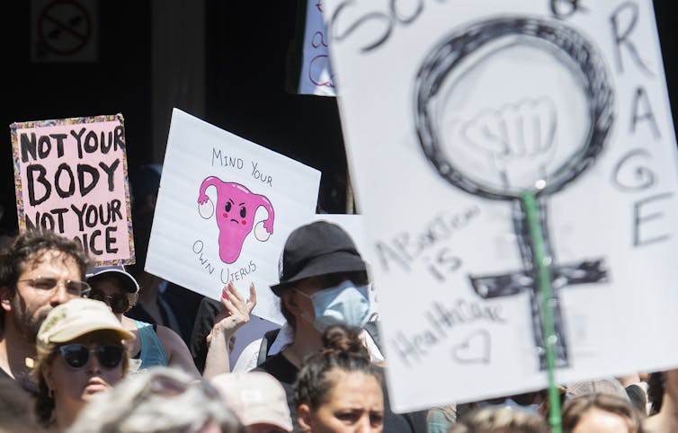 People carry signs at a protest. One says Not Your Body Not Your Choice, another says Mind Your Own Uterus.