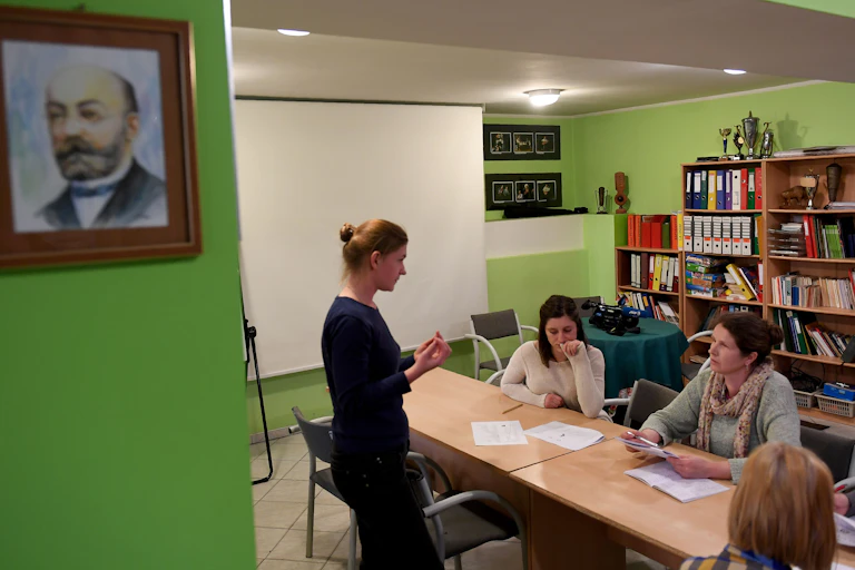 A person stands in front of a few people gathered around a table, with a painting of a man's head on a wall nearby