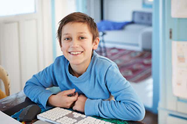 A young boy in a blue shirt smiles as he works at his computer.