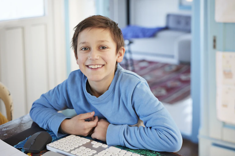 A young boy in a blue shirt smiles as he works at his computer.