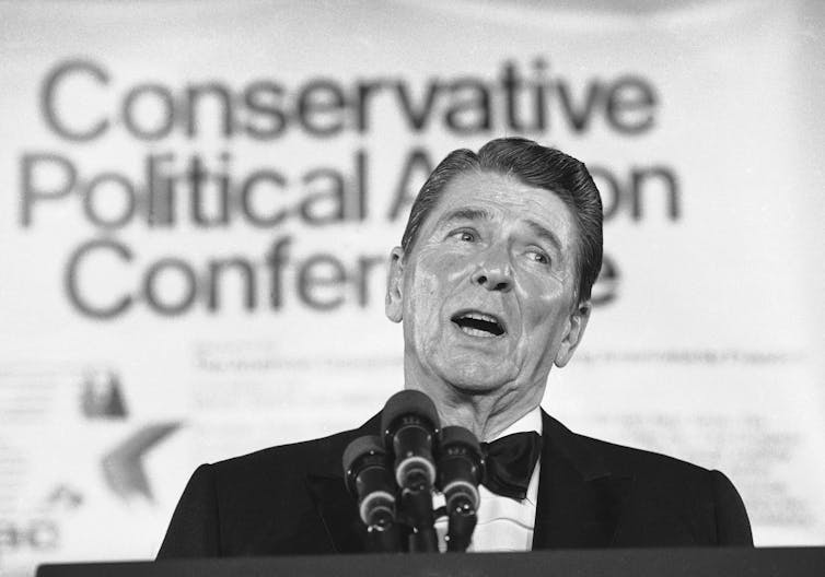Ronald Reagan, wearing a black tie and black jacket, speaking to an audience.