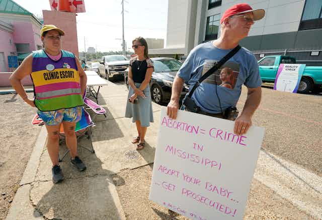 A man wearing red hat holds a sign that reads ‘abortion = crime’ as a woman wearing a sign that says clinic escort stand in front of an abortion clinic
