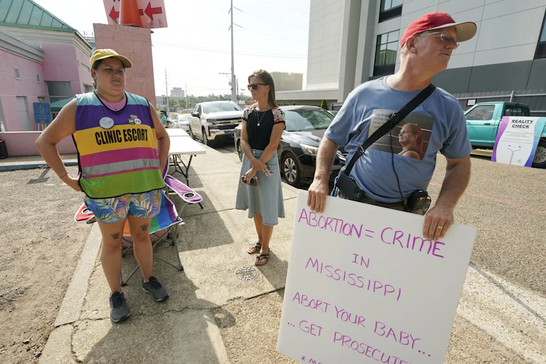 A man wearing red hat holds a sign that reads ‘abortion = crime’ as a woman wearing a sign that says clinic escort stand in front of an abortion clinic
