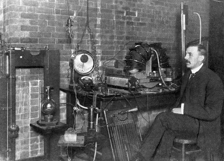 Black and white photo of man in old style dress sitting in front of an elaborate contraption.