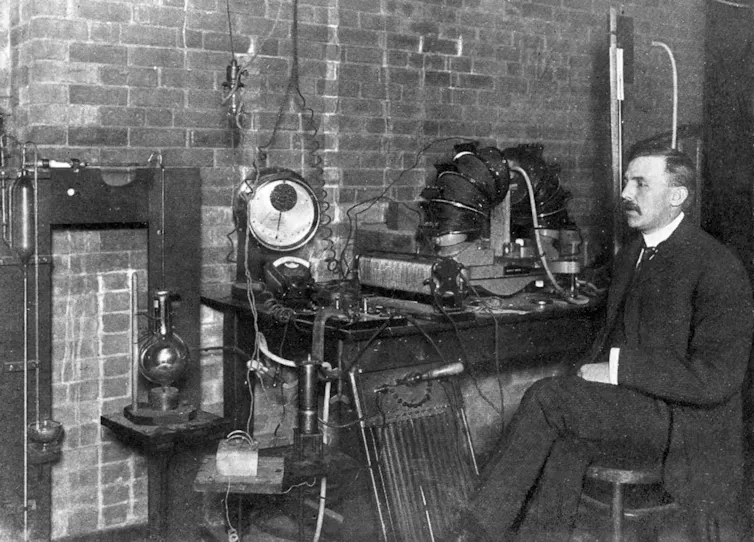 Black and white photo of man in old style dress sitting in front of an elaborate contraption.
