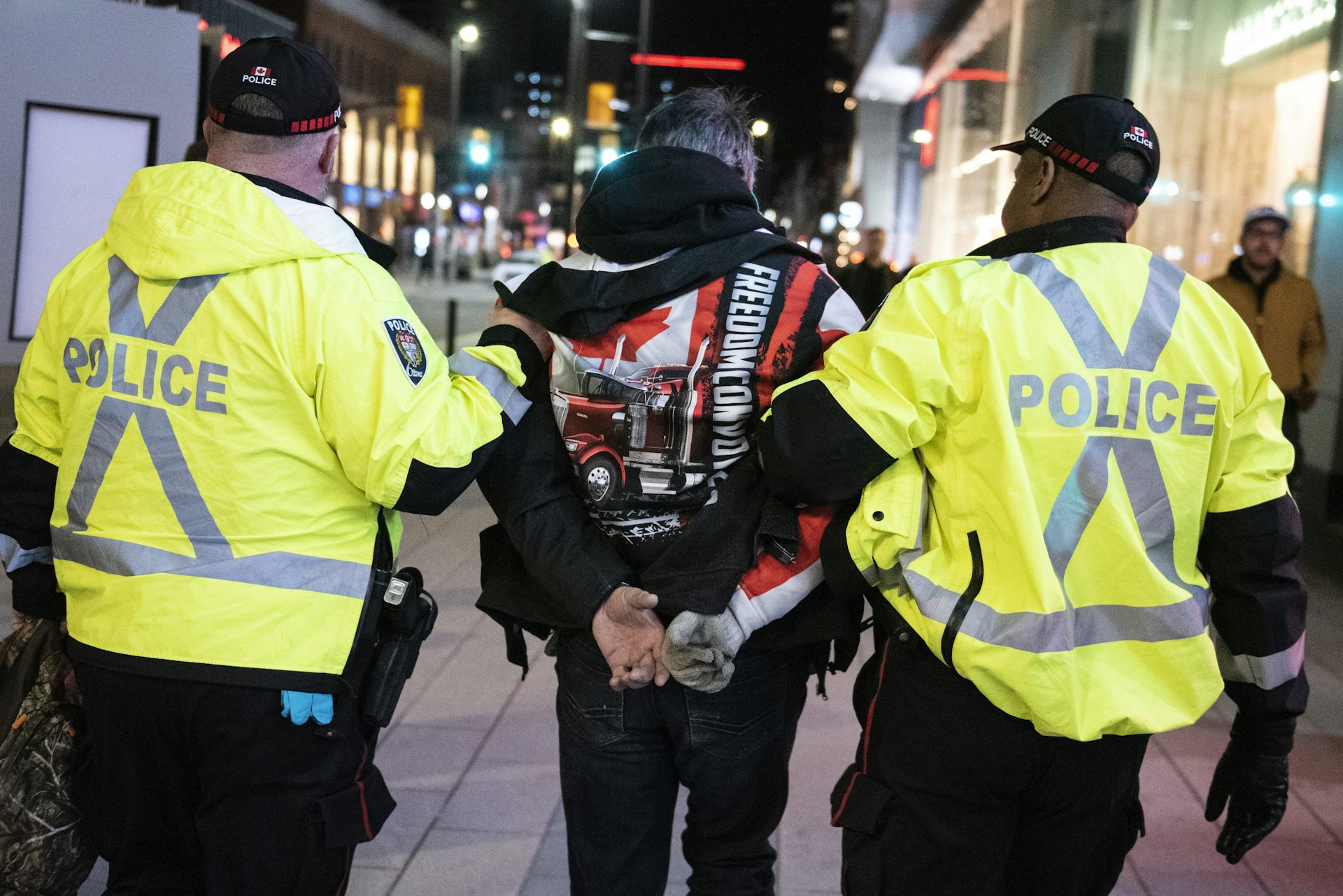 A person in handcuffs wearing a freedom convoy jacket held by police in yellow vests.