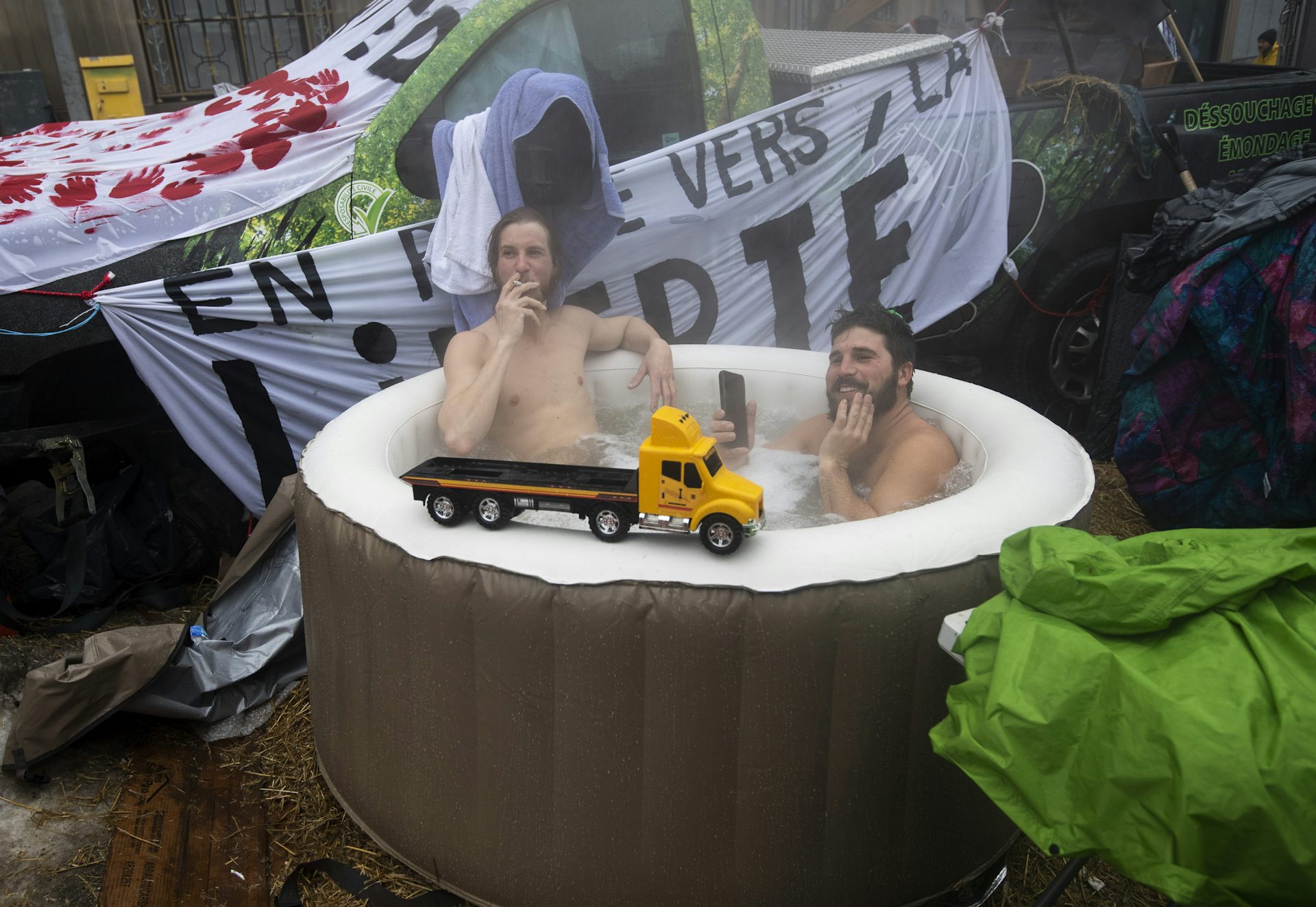Two bare-chested men sit in a hot tub. One smokes a cigarette.