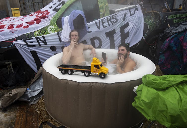 Two bare-chested men sit in a hot tub. One smokes a cigarette.