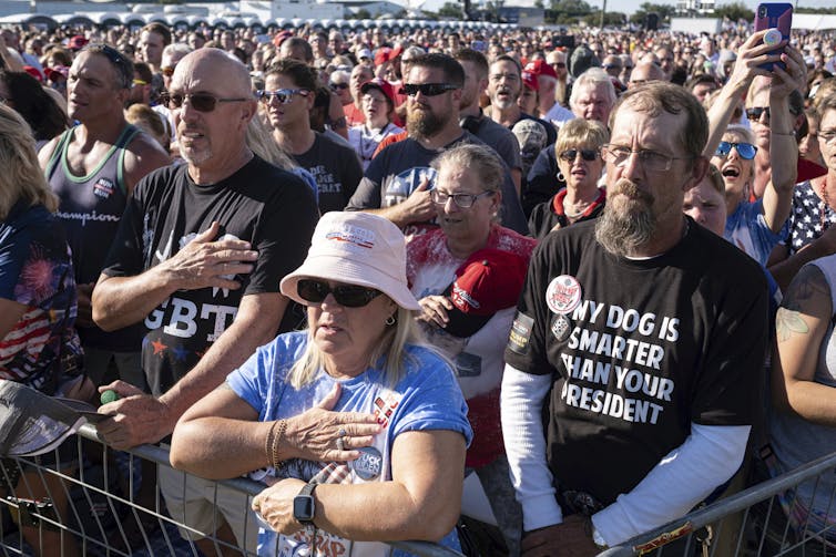 Trump supporters put their hands on their hearts as they sing the American national anthem at an outdoor Trump rally.