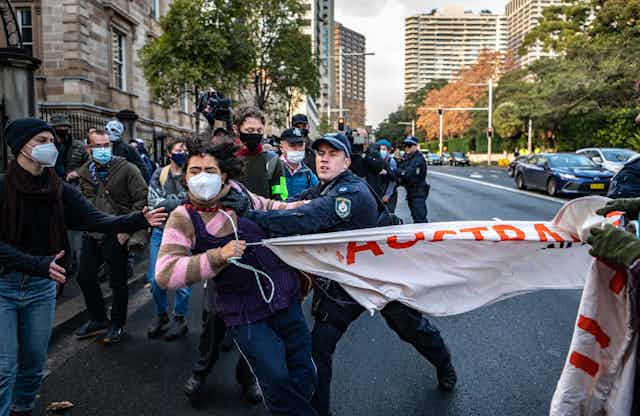 A police officer pushing a masked protester holding a banner