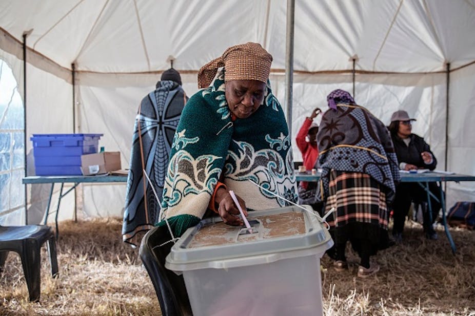 A woman wearing a blanket and a turban puts a ballot paper in a ballot box.