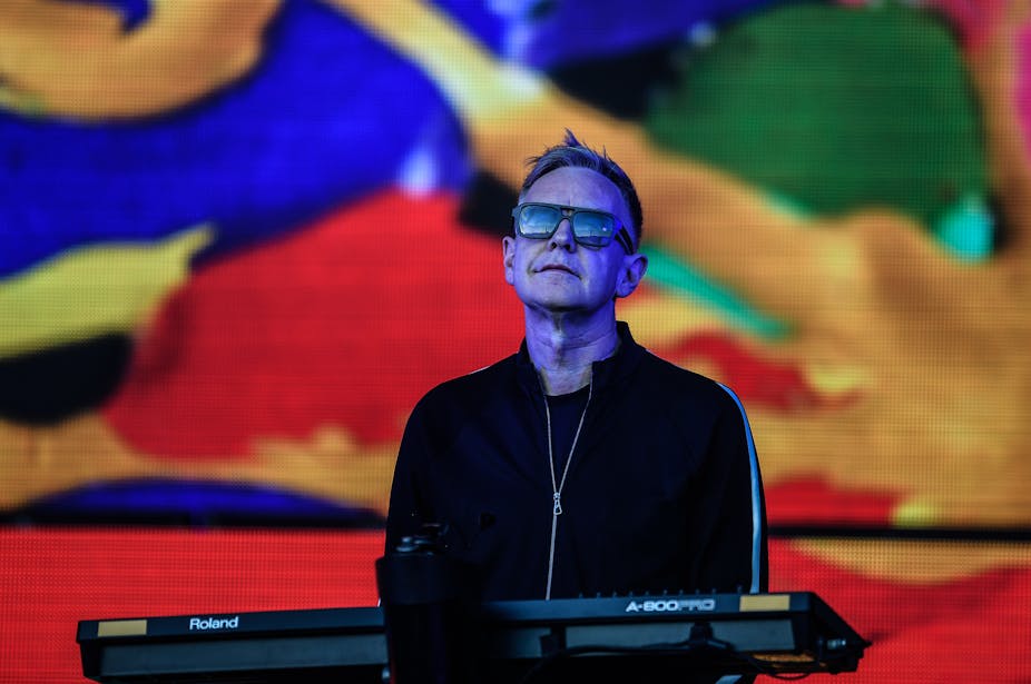 Andrew Fletcher of Depeche Mode stands on stage behind his keyboard wearing sunglasses.
