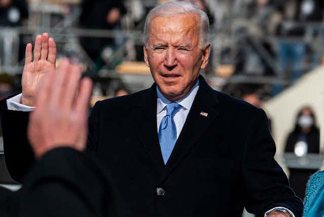 Joe Biden with his right hand raised, squinting into the sunlight as he is sworn in.