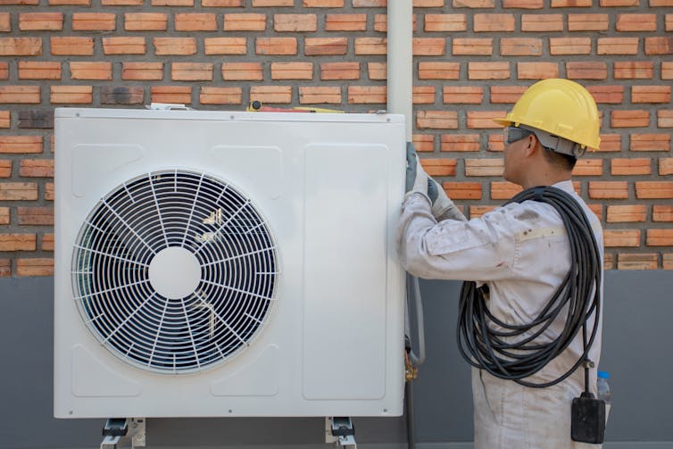 An engineer adjusts the external fan unit of a heat pump on the side of a house.
