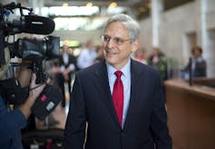 A grey-haired man wearing glasses in a suit with a red tie smiles as he walks past reporters.