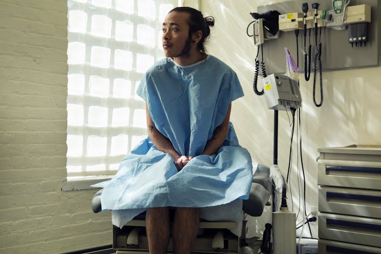 A genderqueer person sitting in a hospital gown sitting in an exam room