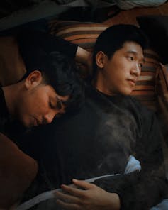 A person who presents as male lying with one hand behind his head, looking off to the side, and another person asleep with his head on his chest.