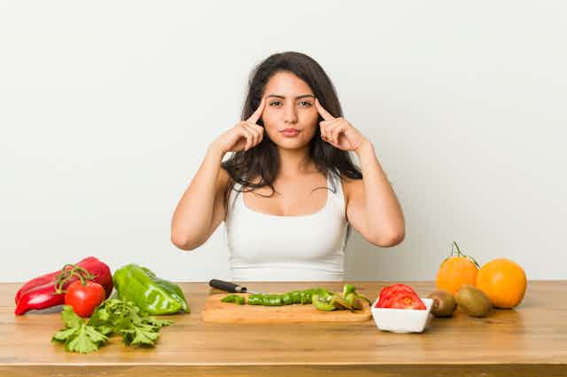Una mujer ante varios platos de fruta y verdura se señala las sienes con los dedos.