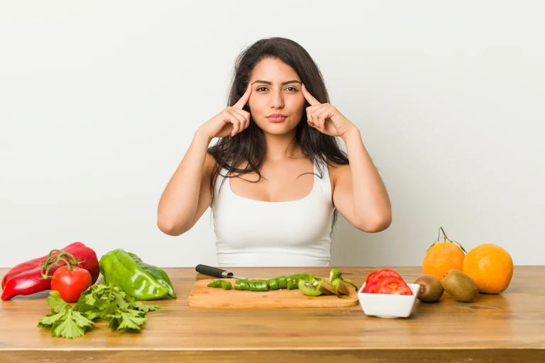 Una mujer ante varios platos de fruta y verdura se señala las sienes con los dedos.