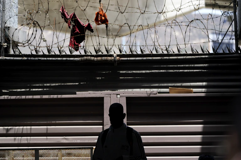The silhouette of a man in shadow in seen in front of a barrier with barbed wire and spikes.