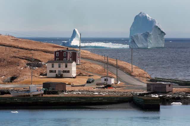 Un point de terre avec quelques bâtiments et des bateaux avec un très grand et allongé iceberg dans l'eau derrière lui. 