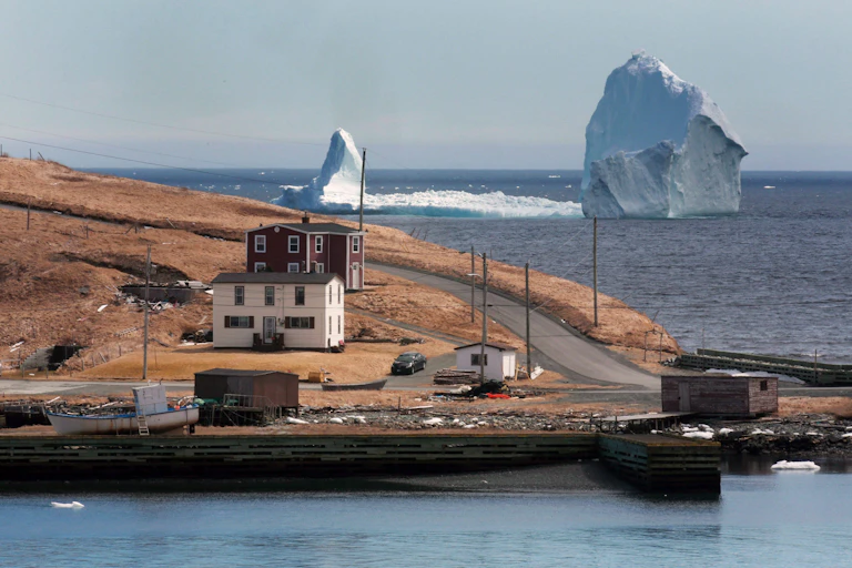 Un point de terre avec quelques bâtiments et des bateaux avec un très grand et allongé iceberg dans l'eau derrière lui.