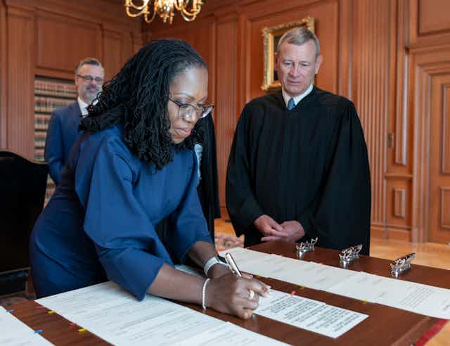 Dressed in a blue dress, Ketanji Brown Jackson signs a piece of paper while Chief Justice John Roberts looks on.
