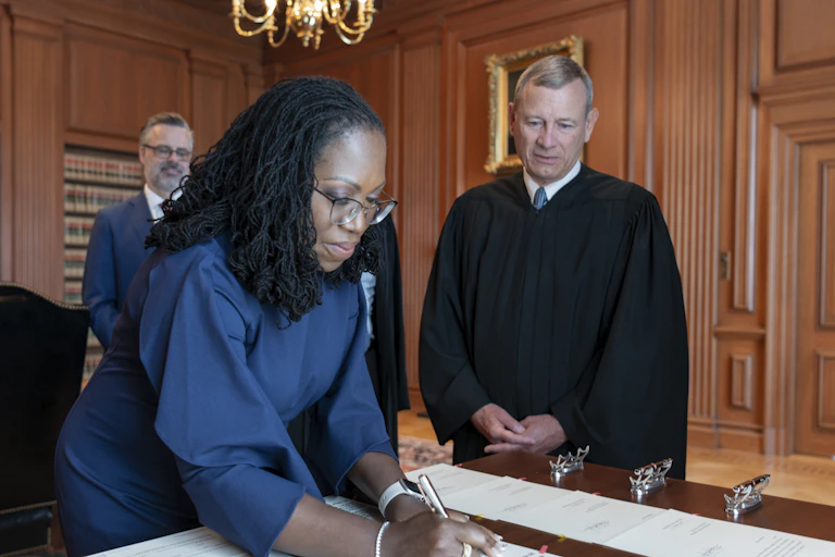 Dressed in a blue dress, Ketanji Brown Jackson signs a piece of paper while Chief Justice John Roberts looks on.