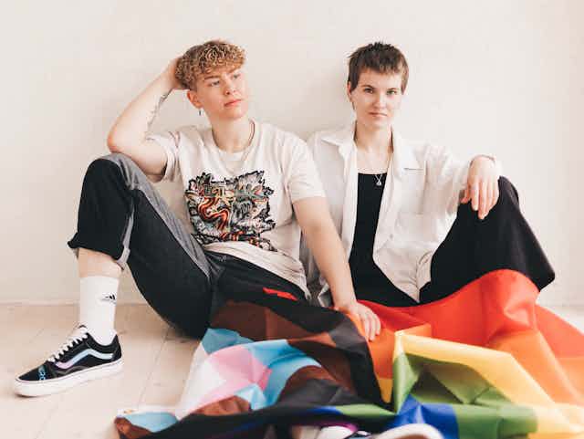Two young people in casual clothes sitting on the floor with a trans flag and a rainbow Pride flag