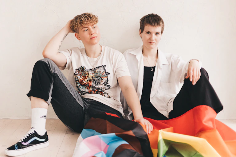 Two young people in casual clothes sitting on the floor with a trans flag and a rainbow Pride flag