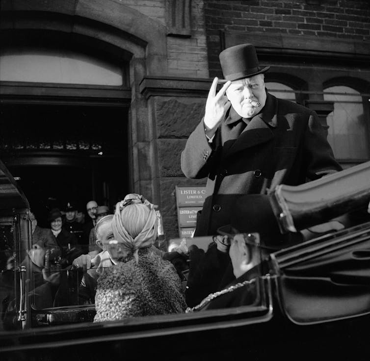 Black and white photo of Winston Churchill in an open-top car, wearing a top hat and with a cigar in his mouth, giving the v for victory sign