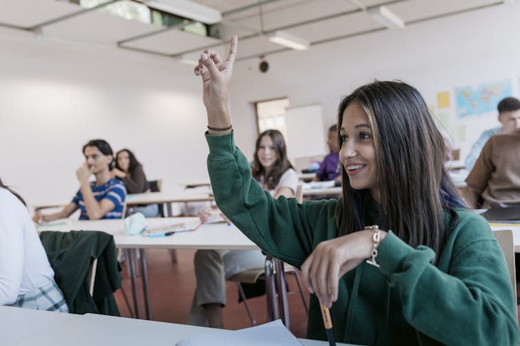A girl student in a green sweatshirt raises her hand in a class full of students