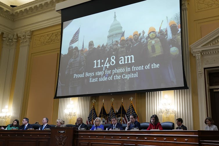 People sit in a row behind a dais and behind them a large screen shows an image of a crowd in front of the Capitol
