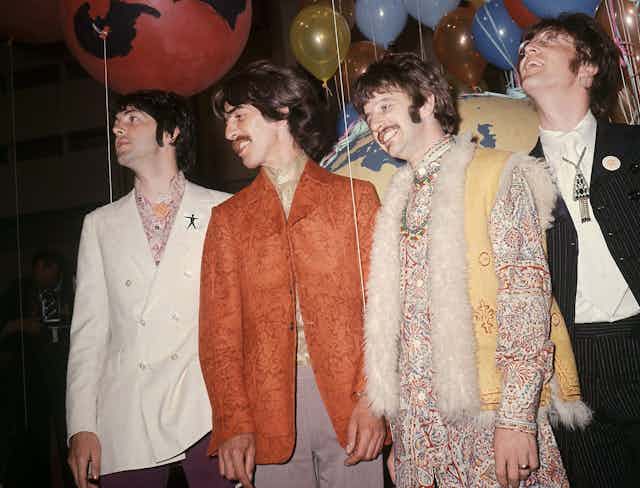 Four men seen standing in snazzy outfits, a white jacket, a orange jacket, a fur-trimmed vest and a black jacket, amid balloons. 