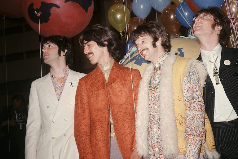 Four men seen standing in snazzy outfits, a white jacket, a orange jacket, a fur-trimmed vest and a black jacket, amid balloons.