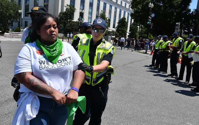 A black woman wearing a teeshirt that says 'We don't back down' is detained by a police officer.
