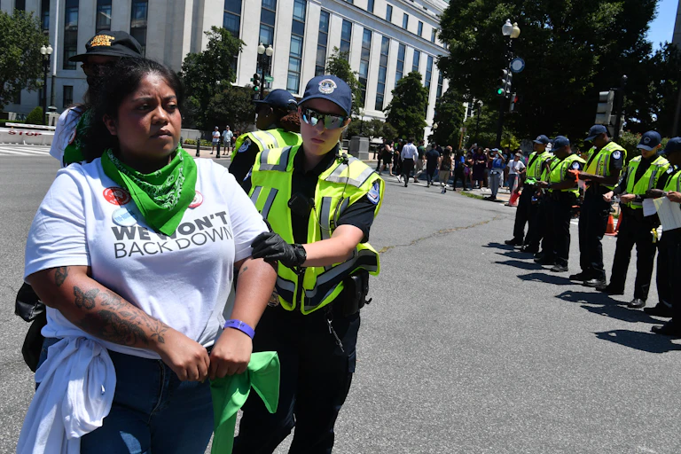 A black woman wearing a teeshirt that says 'We don't back down' is detained by a police officer.