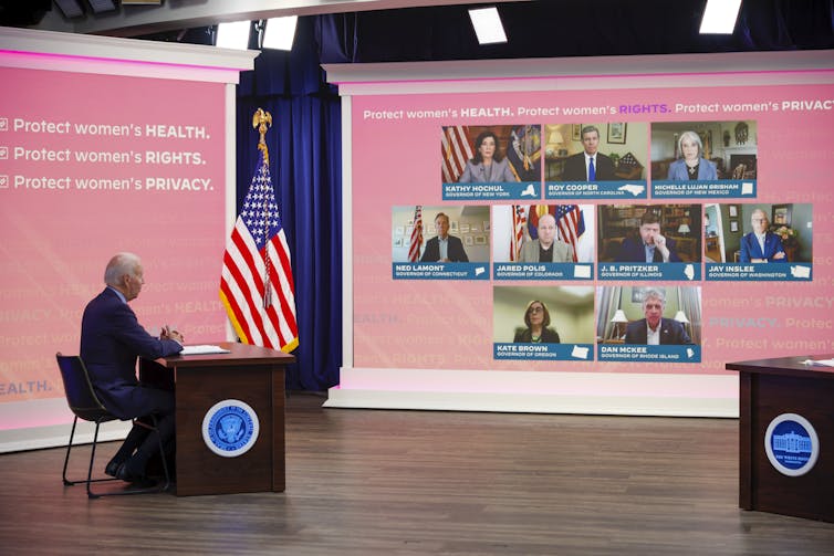 An elderly white man sits behind a desk while holding a virtual meeting showing participants on a large pink screen.