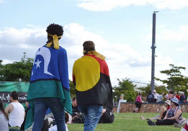 Two people walk around a festival. One person has the Torres Strait Islander flag around their shoulders. The other person has an Aboriginal flag draped around their shoulders.