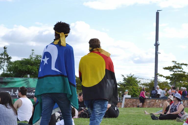 Two people walk around a festival. One person has the Torres Strait Islander flag around their shoulders. The other person has an Aboriginal flag draped around their shoulders.