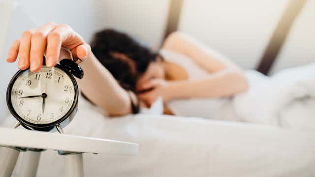 Mujer semidormida en la cama con la mano sobre un despertador negro.